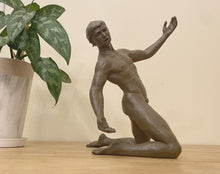 Load image into Gallery viewer, A realistic nude male figurative sculpture in a clay-like brown finish, kneeling on a light wood surface with a potted plant and beige wall in the background.