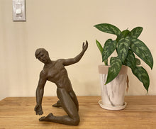 Load image into Gallery viewer, A dark bronze sculpture of a kneeling nude male dancer figure posed next to a potted green plant on a wooden tabletop against a beige wall. Light switch at top left is for size comparison.