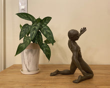 Load image into Gallery viewer, A dark bronze sculpture of a kneeling male dancer with one arm raised, placed on a wooden table next to a potted green plant in a white marble-patterned pot against a beige wall.