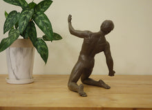 Load image into Gallery viewer, A dark brown sculptural figurine of a kneeling nude human figure with left arm raised, displayed on a light wood tabletop next to a potted green plant against a plain beige wall. See the muscular back of the male dancer in solid bronze.