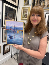 將圖片載入圖庫檢視器 A woman with shoulder-length light brown hair stands in an art gallery holding a framed landscape painting of a harbor scene with boats. She is wearing a gray pleated top and a pearl necklace.