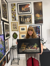 Cargar imagen en el visor de la galería, The female artist Kelly Borsheim is holding a framed still life painting she made in a gallery room with many other of her framed artworks on the walls and pedestals behind her.