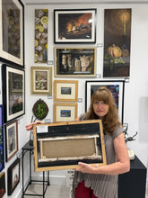 Cargar imagen en el visor de la galería, A woman stands in a gallery room holding up the back of a framed artwork. The room has many framed paintings and prints on the walls, and the woman is holding a large framed piece with a light wood border and dark painted canvas wrapped over stretcher bars and placed inside the frame.