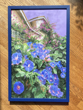 將圖片載入圖庫檢視器 Blue wood framed original painting of blue morning glories growing abundantly on the street level of a house with pink bougainvillea up on the balconies, all under a purple sky. Painting resting on a wood floor in the sunlight.