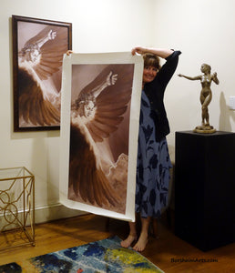 Artist Kelly Borsheim holds up the 50-inch tall print of her Icarus painting up against the length of her body.  The framed 40-inch tall version hangs on the wall behind her.  To the right is Kelly's sculpture "Sirenetta" aka The Little Mermaid nude woman sculpture.