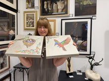 Charger l'image dans la galerie, A woman holding two square canvas prints featuring colorful bird illustrations, standing in an art gallery with framed paintings on the wall behind her. She is showing how thick the wrapped canvas sides are, as well as her signature on the bottom left of each half of the diptych about Painted Bunting birds.