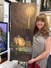 將圖片載入圖庫檢視器 A woman stands in an art gallery holding a large vertical canvas painting of a still life scene with a glowing candle, a pumpkin, and greenery on a shelf.