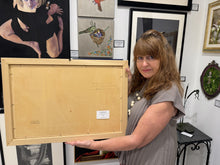 Cargar imagen en el visor de la galería, A woman in a gray dress holds up the back of a large framed artwork painted on a wood panel. The setting is an art gallery or studio with her framed paintings and prints hanging on the walls behind her.