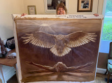 Cargar imagen en el visor de la galería, A woman holds up a large painted canvas featuring a glowing owl with spread wings over a reclining figure, photographed indoors in a home setting with framed pictures on the wall behind her.