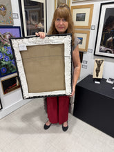 Charger l'image dans la galerie, A woman in a gallery room holds up the back of a framed artwork, showing the canvas, wide unpainted border, and handwritten label. The room has other framed art pieces on the walls and a black display table.