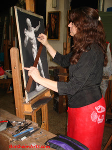 Artist Kelly Borsheim uses a mahl stick to put some subtle details in the highlight areas of the body of the Écorché archer.  She stands in front of an art studio easel in a black top and a red sarong worn as a skirt.