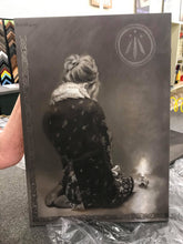 Laden Sie das Bild in den Galerie-Viewer, This is the left portion of the monochromatic diptych oil painting Luminosity, which depicts a figure of a young woman who sits on her legs in a winter coat.  She is lost in her own thoughts as she looks towards the flame of a single candle resting on the floor beside her.