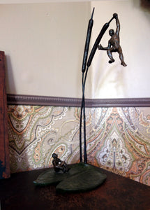 Sculpture of two tiny bronze men on a bronze lily pad and cattails.  One man dangles precariously while the other one sits looking up at him.  Shown here in the corner of a desk in a vintage decor home.