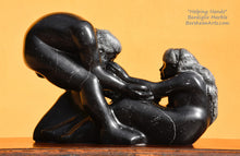 Load image into Gallery viewer, reflections Helping Hands by Kelly Borsheim Couple Art Carved from a Marquina black marble / limestone from Spain, this sculpture depicts a man bending over forward to help a seated woman stand up. Her hands reach up towards his bearded face, but it is the moment before she is close enough to reach him.  Woman on the right, yellow wall in the background