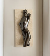 A bronze-finished sculpture of a nude female figure mounted on a rectangular beige stone tile, displayed against a neutral beige wall.
