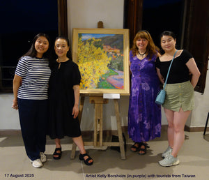 Four women posing in front of a colorful landscape painting on a wooden easel, artist in a purple dress standing out.