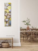 Load image into Gallery viewer, vertical painting of lemons and pastries is hung in the entry way to a dining room in neutral colors with wicker furniture 
art by Kelly Borsheim
