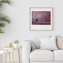 Load image into Gallery viewer, This print of a pink wall in Morocco and a shy boy wearing white is shown in mock frame and a light neutral colored living room scene with comfortable white couch and side table with plants.
