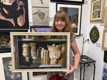 Cargar imagen en el visor de la galería, A woman holding a framed painting of a bookshelf scene with books, vases, and a crown, photographed inside an art gallery with other paintings on the walls.
