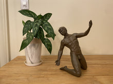 Load image into Gallery viewer, A dark brown tabletop sculptural figurine in solid bronze of a kneeling male statue without costume displayed on a wooden table next to a potted green plant against a beige wall.
