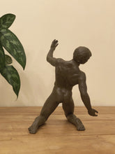 Load image into Gallery viewer, A dark brown matte bronze sculpture of a muscular male figure in a dynamic pose, standing on a light wood surface against a beige wall with a green plant leaf visible on the left.
