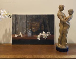 Bronze sculpture of a couple embracing while standing next to a brown and neutral colored still life oil painting on the wooden surface of a dresser.