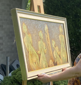 Framed artwork of abstract figures held at an angle on an easel by a person outdoors.