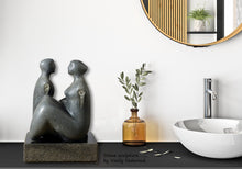 Load image into Gallery viewer, Decorative stone sculpture of a couple seating facing one another on a shelf with a plant and sink in the background
