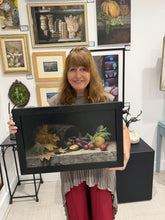 Cargar imagen en el visor de la galería, A woman artist holding a framed still life painting featuring autumn leaves, a dark barrel, and small red radishes, with a gallery wall of other framed art behind her.
