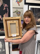 將圖片載入圖庫檢視器 A woman in a gray pleated dress holds up a light natural wood picture frame showing the back side with mounting material and a small label. She is standing in an art gallery or studio with framed paintings and prints hanging on the wall behind her.
