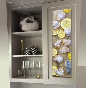 tall thin painting of Italian pastries with lemon slices hangs on a grey cabinet in a modern style kitchen.  The shelves to the left hold a sculpture, an arty teapot and cup, candle holder, empty... lower shelf has clean empty wine glasses