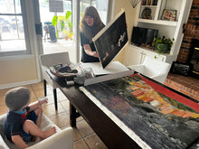將圖片載入圖庫檢視器 Woman artist shows child nephew how to re-stretch a painted canvas.  There is another large painting on a dining table in a home setting.
