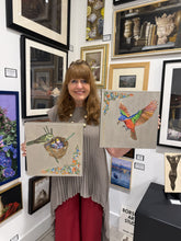 Charger l'image dans la galerie, A woman with shoulder-length brown hair stands in an art studio holding two framed paintings. The left painting shows a green mamma bird in a nest with blue chicks, and the right painting shows a colorful male pappa bird in flight with a worm in beek to feed his young.
