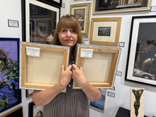 Charger l'image dans la galerie, A woman in a gray striped top holds up two canvases showing the back sides with white labels, standing in an art gallery with many of her framed paintings on the wall behind her.
