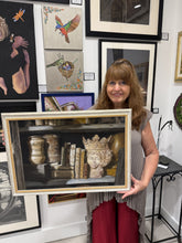 Cargar imagen en el visor de la galería, A woman in a gray sleeveless top and burgundy pants holds up a framed painting of a shelf scene with books, jars, and a decorative bust. The background shows a gallery wall with other framed art.

