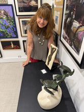 Laden Sie das Bild in den Galerie-Viewer, A woman with reddish-brown hair stands behind a low pedestal in an art gallery. It displays a bronze sculpture of two turtles on a light stone base, with framed paintings hanging on the white walls behind her.
