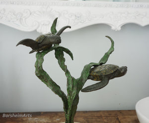 Bronze sculpture of two turtles swimming through three strands of sea kelp with a decorative white frame in the background.