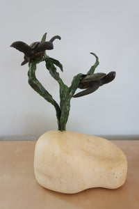 Decorative sculpture in bronze of two sea turtles swimming near a kelp plant growing out of a stone base [as a sandy seabed] on a plain background.