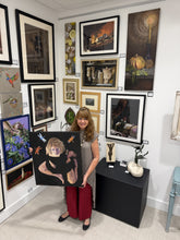 Charger l'image dans la galerie, A woman stands in a gallery room holding a canvas painting of a woman's head and limbs emerging from a solid dark background and dragonflies circling around above her in the air. The background is filled with other framed art pieces on white walls.
