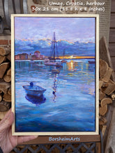 Load image into Gallery viewer, Artist&#39;s hand holding a small vertical painting of boats in a harbor at sunset.  A simple thin light wood frame comes with the acrylic painting on canvas by Kelly Borsheim.
