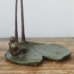 The second man in this original bronze sculpture titled "The Unwritten Future" is sitting on a large lily pad.  He has his hands gripping his legs to balance his body as he looks up to the swinging figure of the other man. Small tabletop sculpture detail image