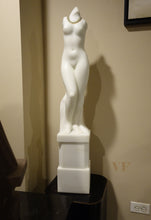 Laden Sie das Bild in den Galerie-Viewer, White marble sculpture of a nude female figure Venus on a pedestal against a beige wall.

