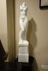 White marble sculpture of a nude female figure Venus on a pedestal against a beige wall.
