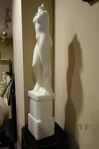 White marble statue of a standing female figure on a pedestal against a beige wall.
