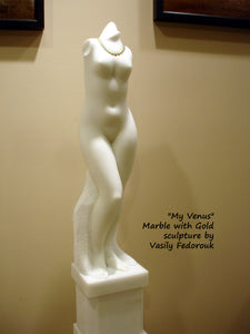 View of the left side of the vertical white marble sculpture of a standing female figure.  You may see here how the shoulder and the neck end (no arms or head).  You may also see some of the pedestal base, part of the same marble piece.  And the gold necklace around the neck.  Art by Vasily Fedorouk