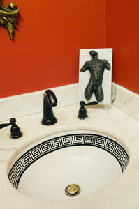 The black patina bronze nude male torso artwork is a dramatic decor addition to this stylish bathroom countertop with black spigot and a sink with a geometric border on the inside edge.  red walls offset the art!  