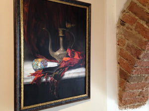 The framed painting, Turkish Light, a still life inspired by the Mediterranean and Turkey, with red and orange scarf and a white marble slab, shown here at art exhibit in Italy, next to a brick arch in the wall.